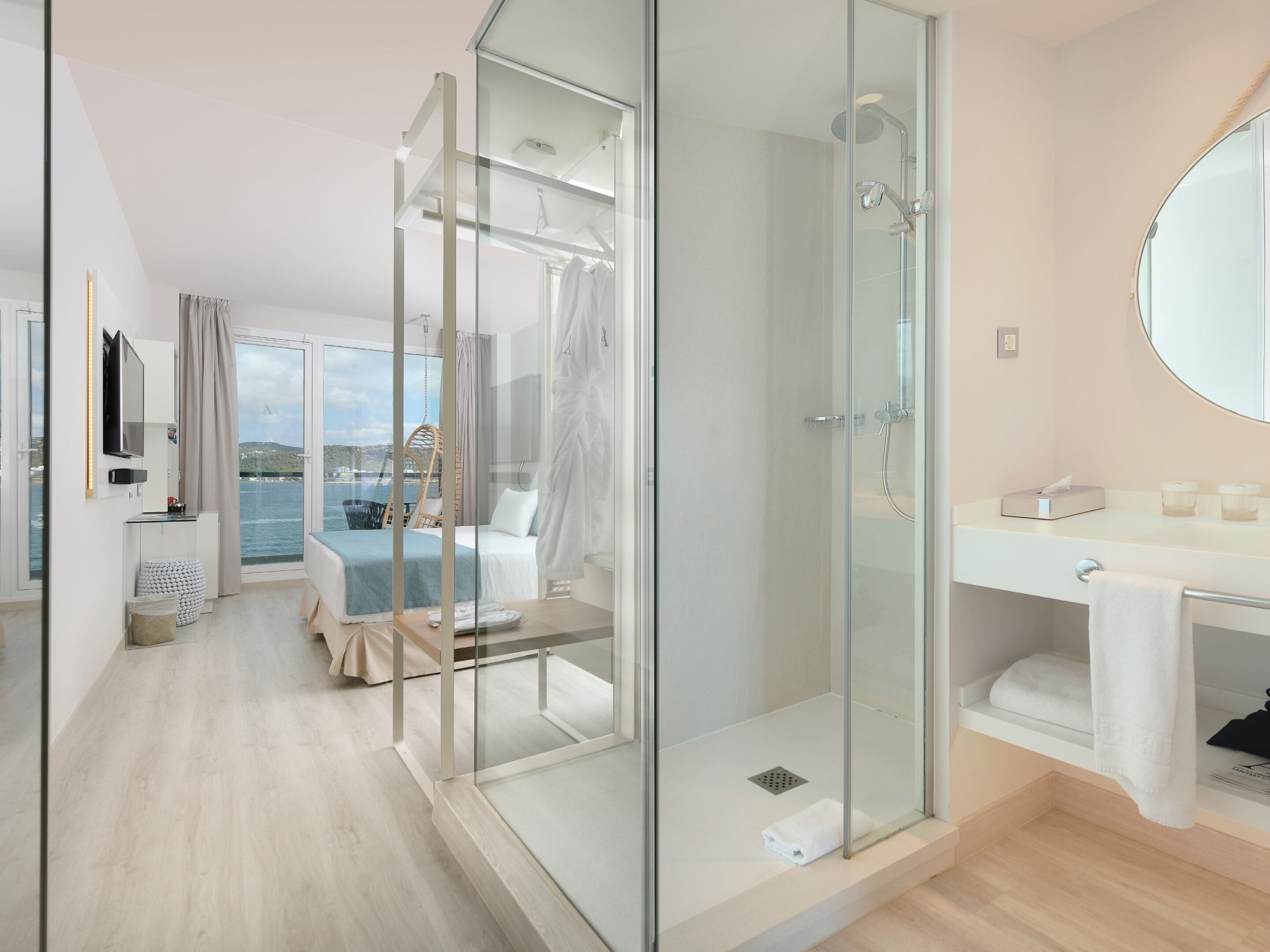 A modern hotel room with a glass-enclosed shower, a bed with a view of the sea, a balcony, and a large mirror.