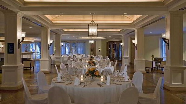 Une salle de banquet avec des tables rondes dressées pour un événement formel, agrémentée de lustres et de chaises blanches.