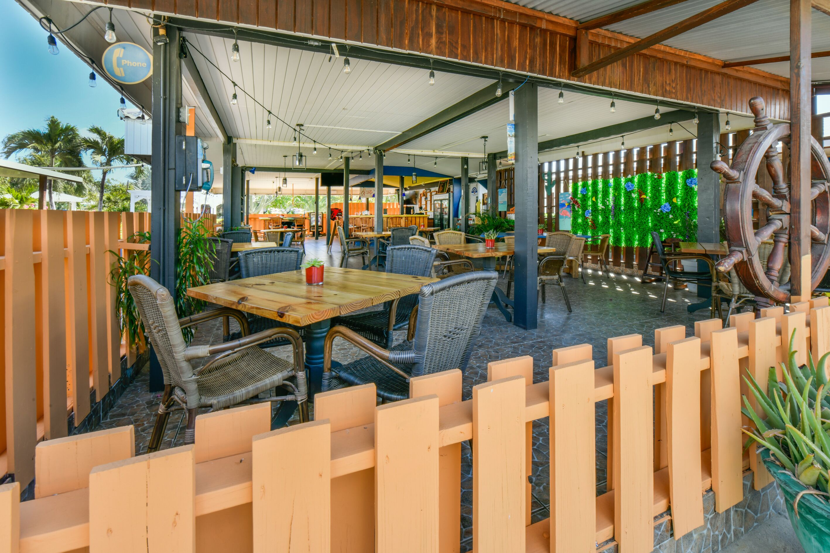 Un área de patio al aire libre con mesas de madera y sillas de mimbre, cerca de madera y una gran decoración de timón de barco.