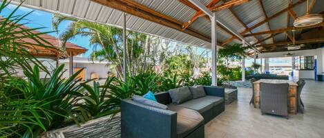 A covered outdoor area with wicker furniture, planters with tropical plants, and a view of the surrounding landscape.