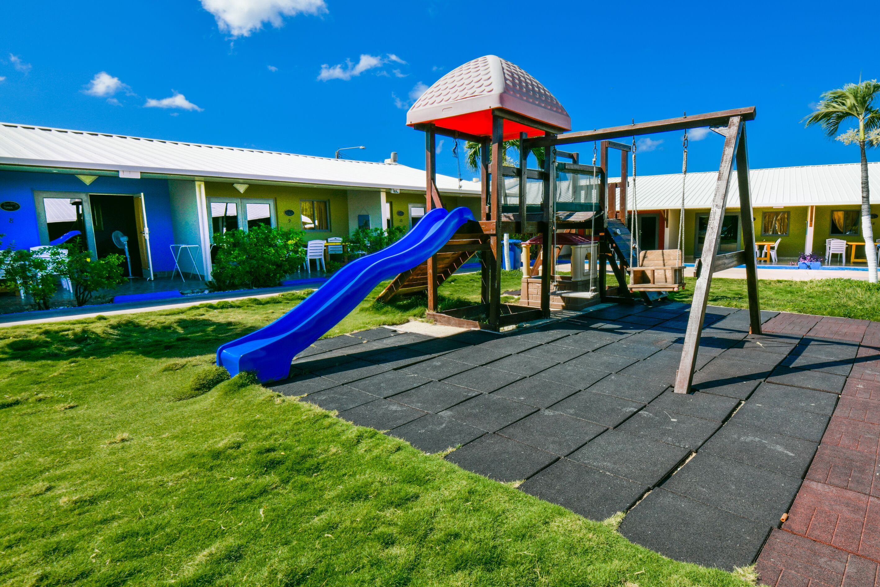 Un parque infantil con un tobogán y una estructura de escalada, rodeado de un césped cuidado y un edificio con techo blanco.