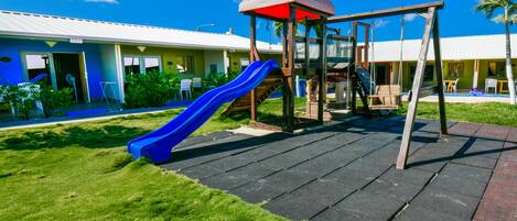 Un parque infantil con un tobogán y una estructura de escalada, rodeado de un césped cuidado y un edificio con techo blanco.