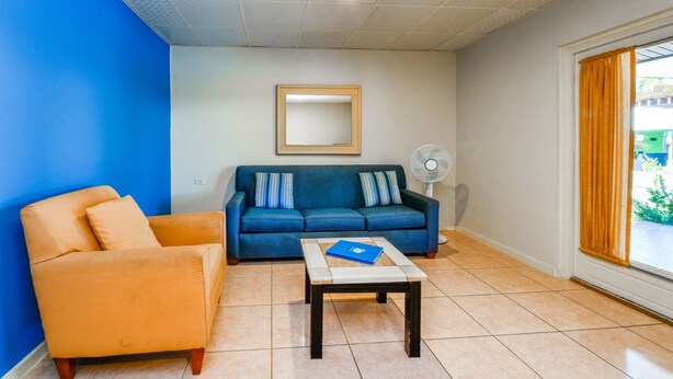 Una sala de estar con un sofá azul, un sillón naranja, una pequeña mesa con una carpeta azul, un ventilador de pie y una ventana con cortinas.