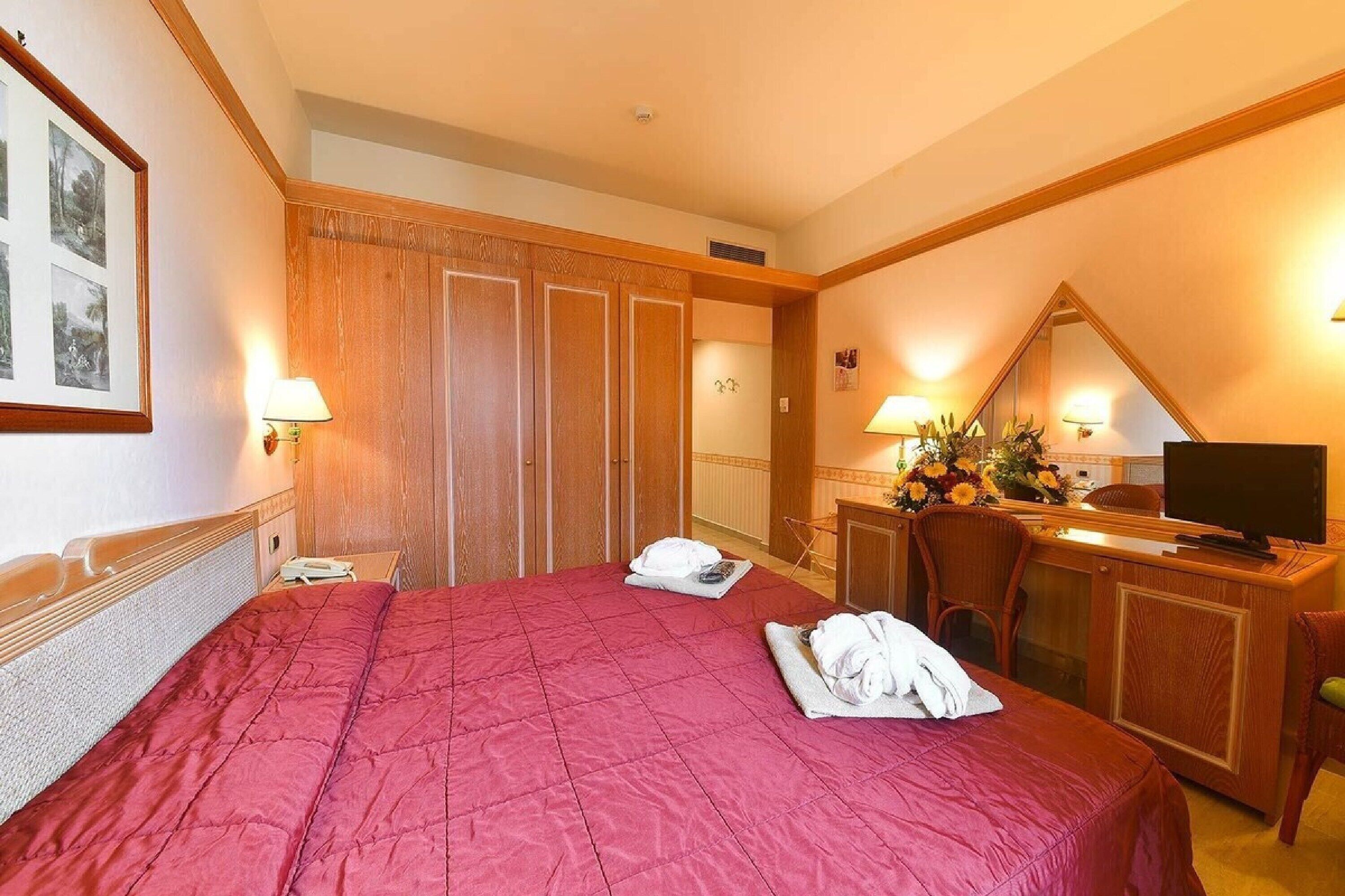 A hotel room with a large bed, a desk with a television, a mirror, and a wardrobe.