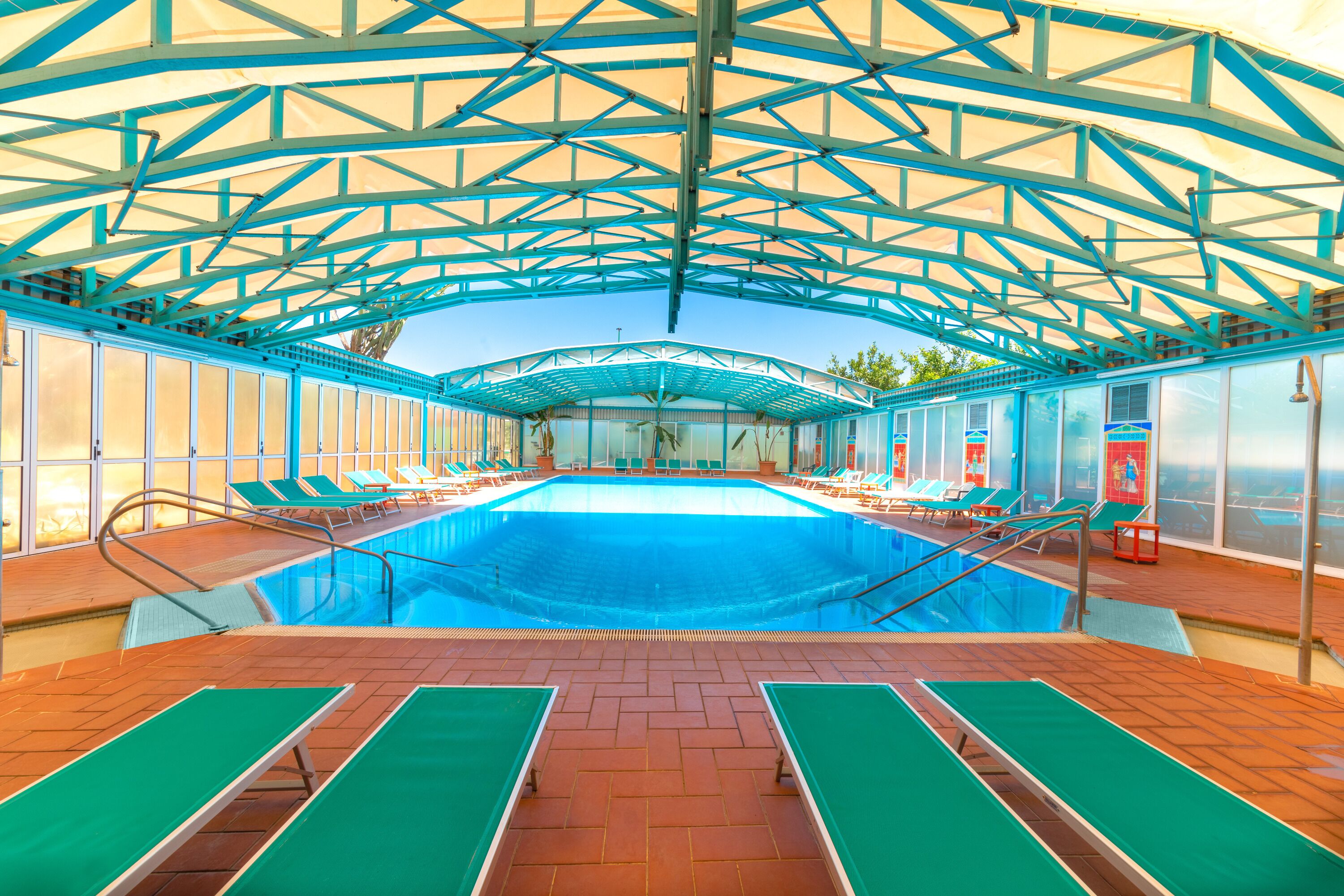 An indoor swimming pool with a blue-tinted roof, tiled floor, and green loungers.