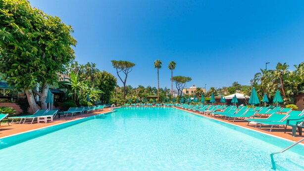 Una piscina rectangular con agua azul cristalina, rodeada de sillas de descanso verdes y sombrillas, con árboles y edificios al fondo.