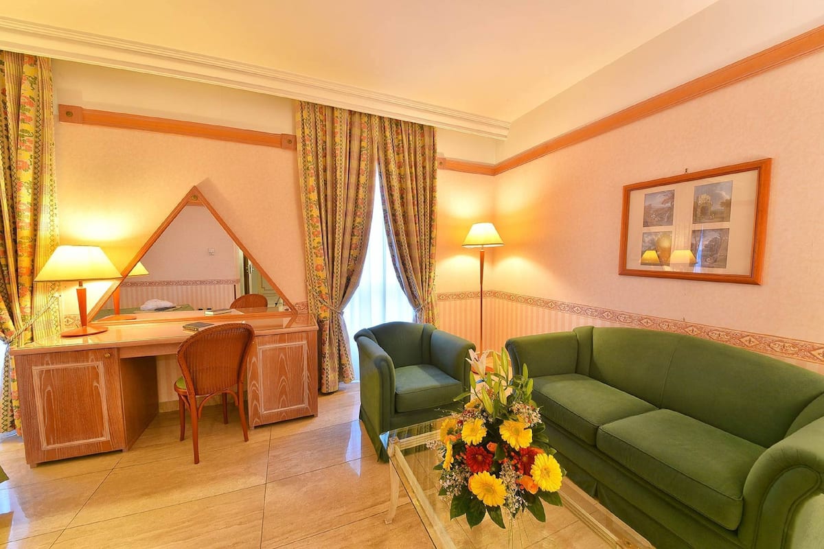 Una camera d'albergo con una scrivania in legno, due sedie, un divano verde, un tavolino in vetro con fiori, uno specchio e due lampade.