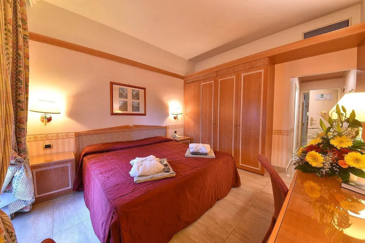 Una camera d'albergo con un letto grande, lampade sui comodini, un armadio in legno e un tavolo da pranzo con un centrotavola floreale.