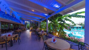 Un área de comedor al aire libre con mesas y sillas, vista a una piscina y un jardín tropical.