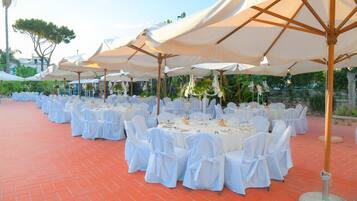 Un área de comedor al aire libre con mesas redondas, manteles blancos y sillas, rodeada de grandes sombrillas.