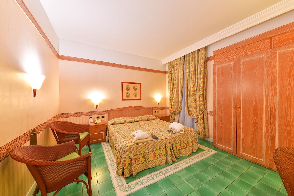 Camera d'albergo con un letto, due sedie, un comodino, un armadio e una lampada a muro.