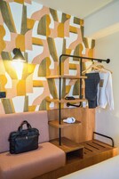 A modern hotel room with a geometric-patterned wall, a wooden bench, a black handbag, and a clothing rack with a white shirt and a dark checkered jacket.