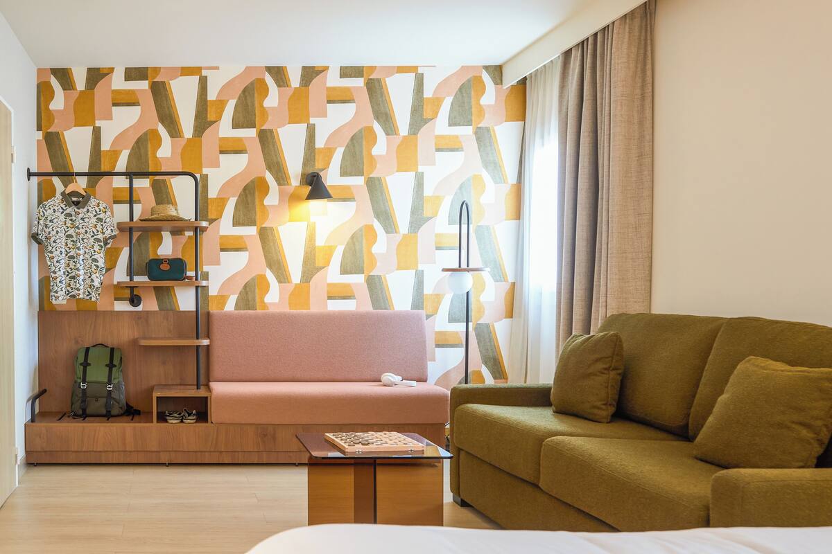 Habitación de hotel moderna con un papel tapiz estampado, un sofá rosa, un sofá verde, una mesita de madera y una pequeña mesa con un tablero de ajedrez.