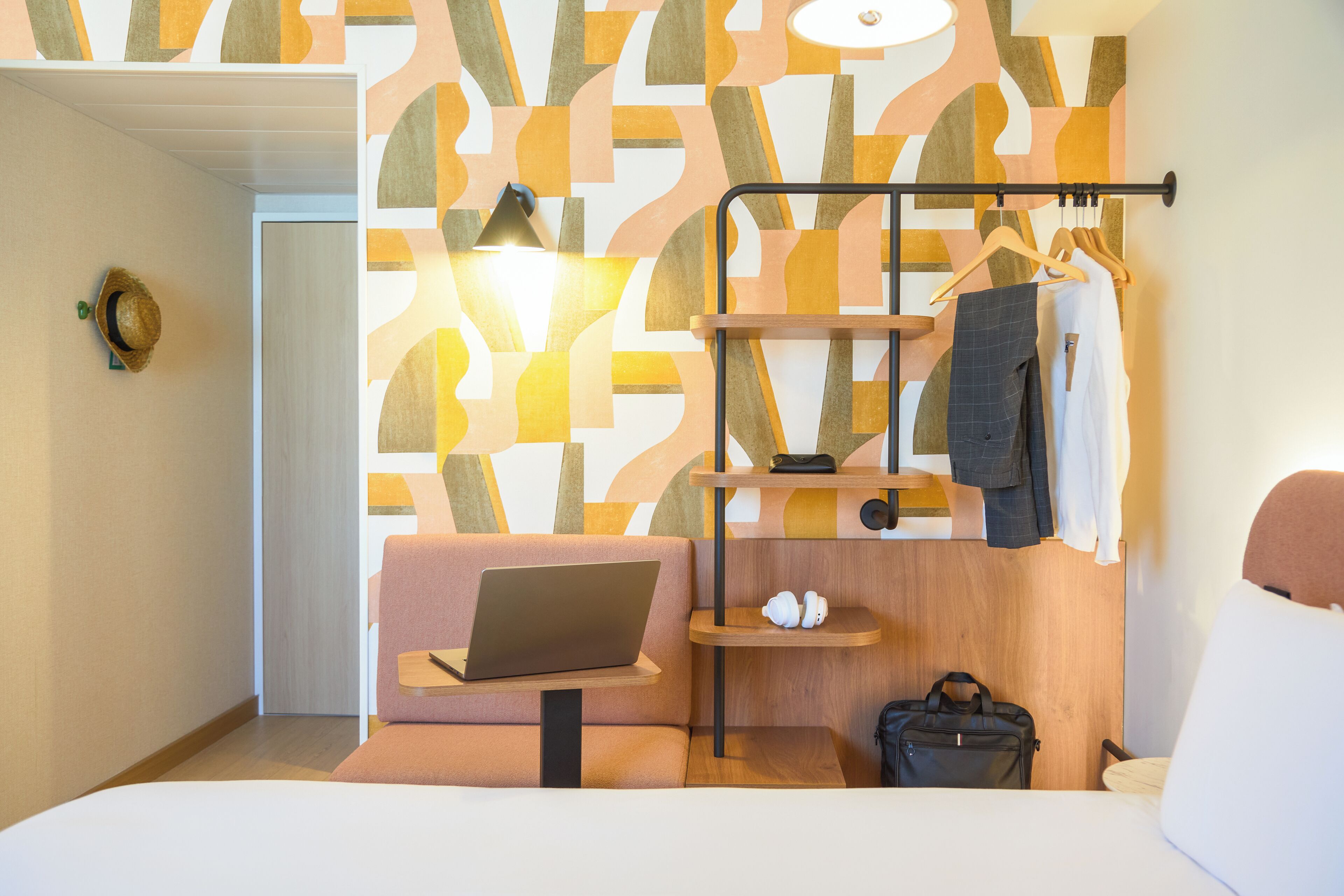 A modern hotel room with a bed, a laptop on a small table, a hanging coat rack, and a wooden shelf with a bag and a pair of slippers.