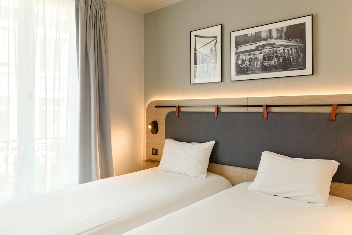 Habitación de hotel con dos camas, un ventanal grande con cortinas translúcidas y dos fotografías en blanco y negro enmarcadas en la pared.