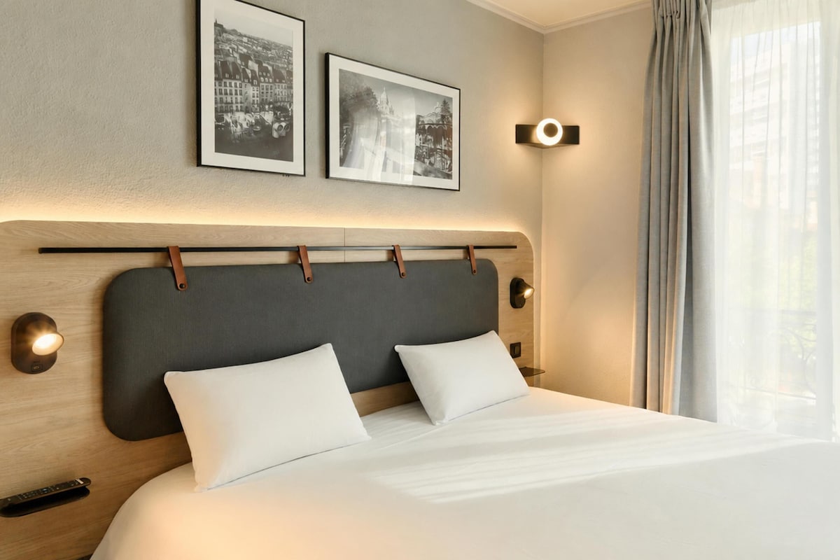 Una habitación de hotel moderna con una cama grande, dos cuadros enmarcados en la pared, una lámpara de noche y una ventana con cortinas.