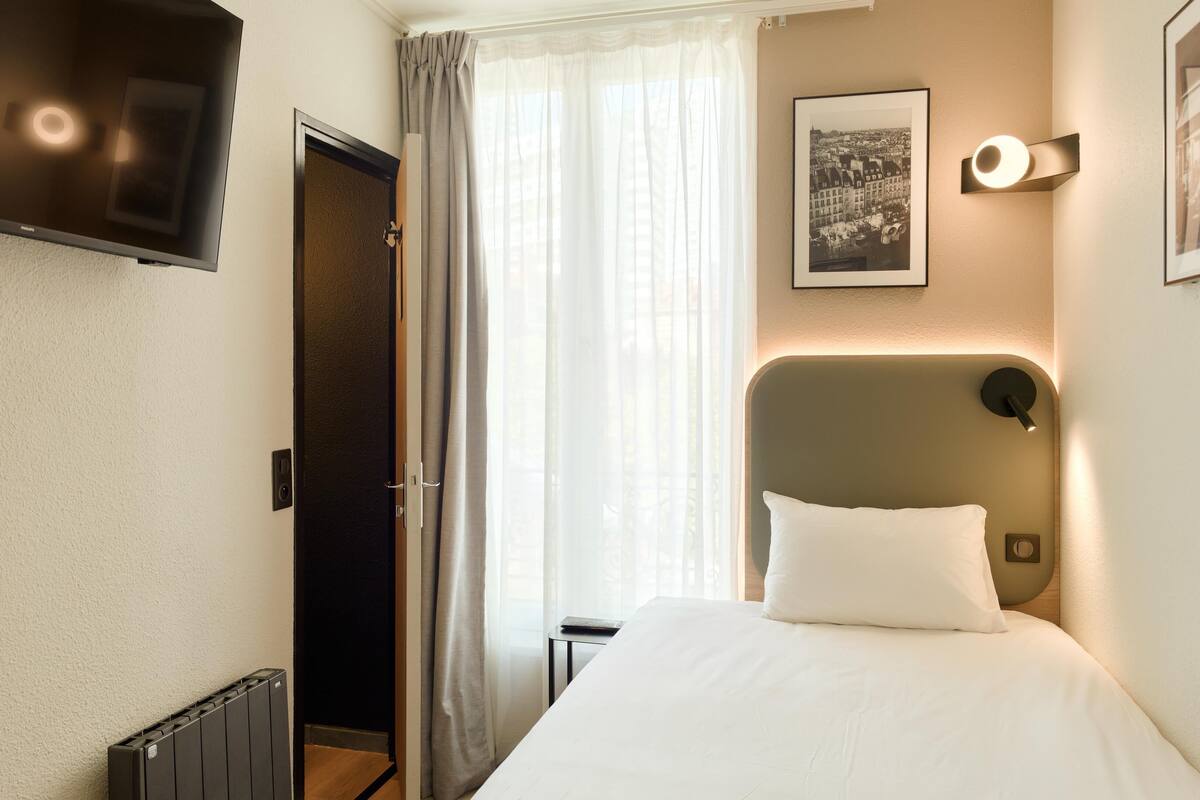Habitación de hotel con cama, televisor montado en la pared, un cuadro enmarcado y una ventana con cortinas.