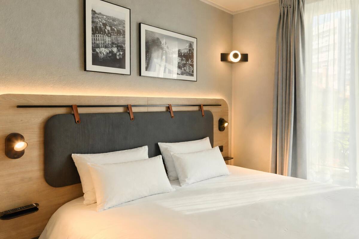 Una habitación de hotel con una cama grande, dos marcos de cuadros en la pared, una mesita de noche con lámpara y una ventana con cortinas.