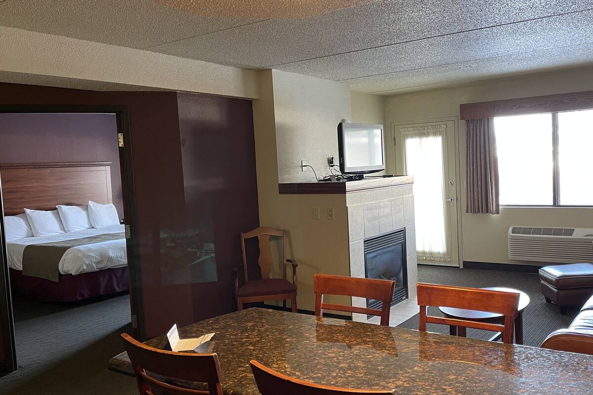 Een hotelkamer met een bed, een eettafel met stoelen, een open haard, een televisie en een raam met gordijnen.
