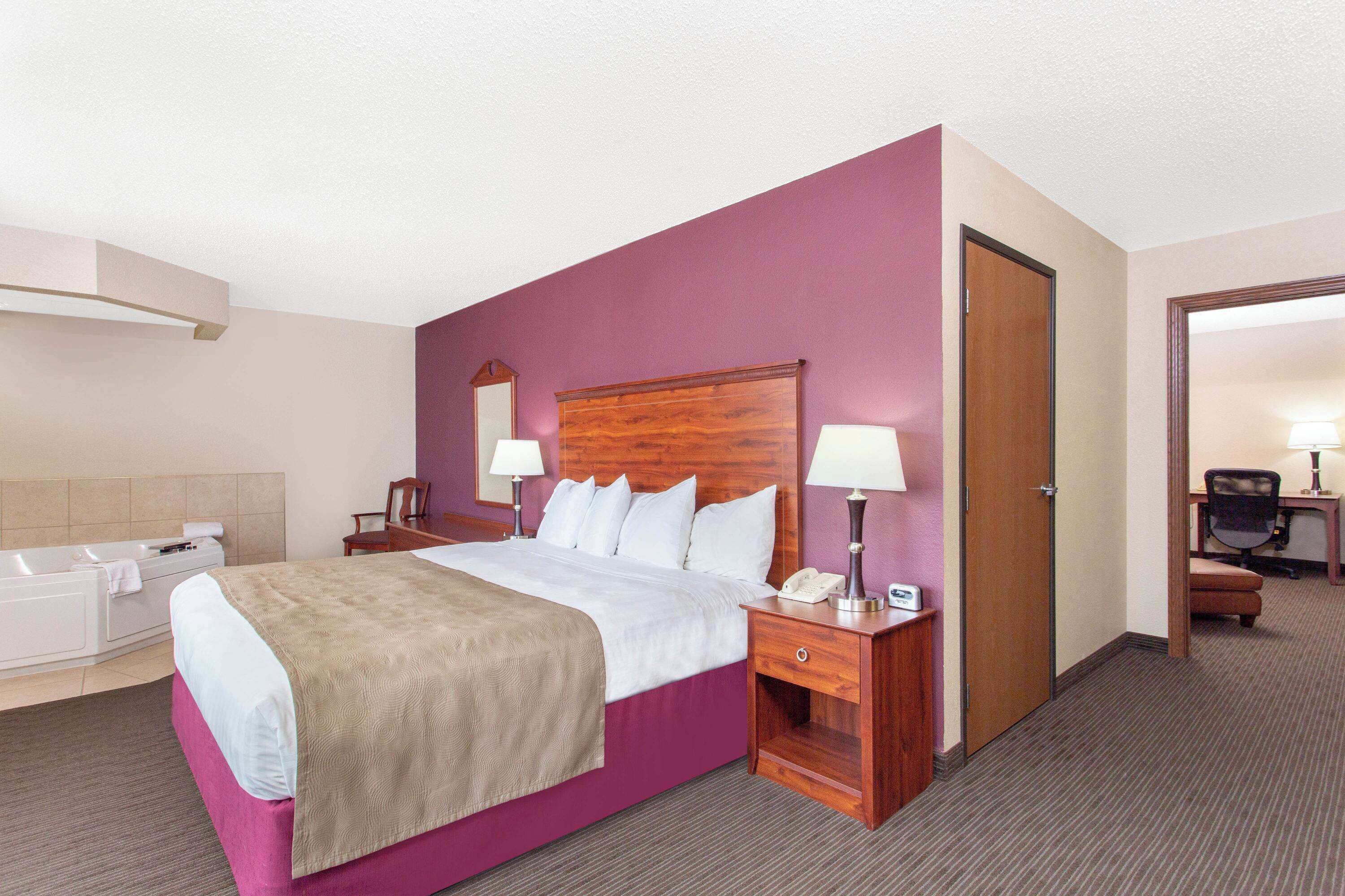 A hotel room with a large bed, a wooden headboard, two bedside tables with lamps, a desk with a chair, and a bathroom with a bathtub and towel rack.