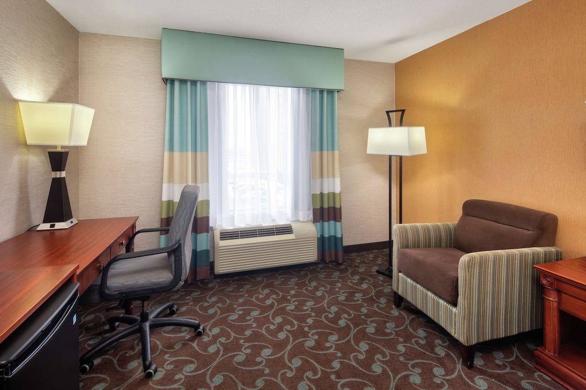 A hotel room with a desk, office chair, armchair, two lamps, and a window with curtains.