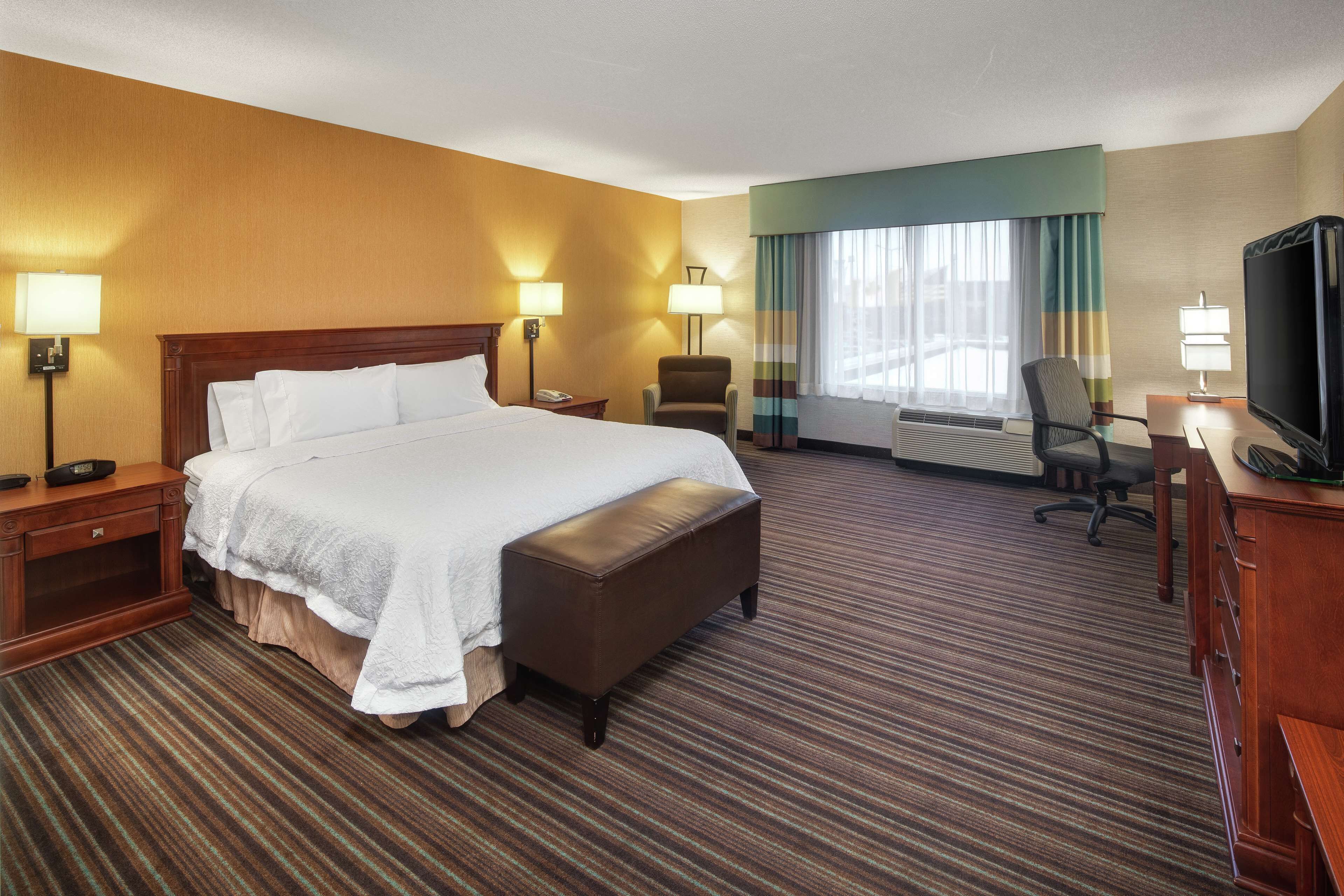Quarto de hotel com uma cama grande, uma escrivaninha com televisão, uma cadeira e uma janela com cortinas.