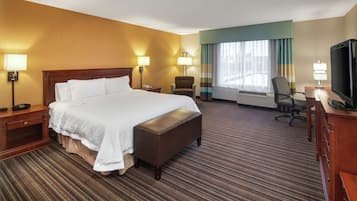 Quarto de hotel com uma cama grande, uma escrivaninha com televisão, uma cadeira e uma janela com cortinas.