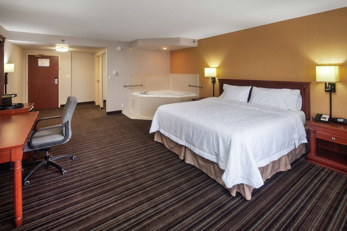 A hotel room with a large bed, a desk, a chair, a desk lamp, a telephone, and a bathroom with a bathtub.