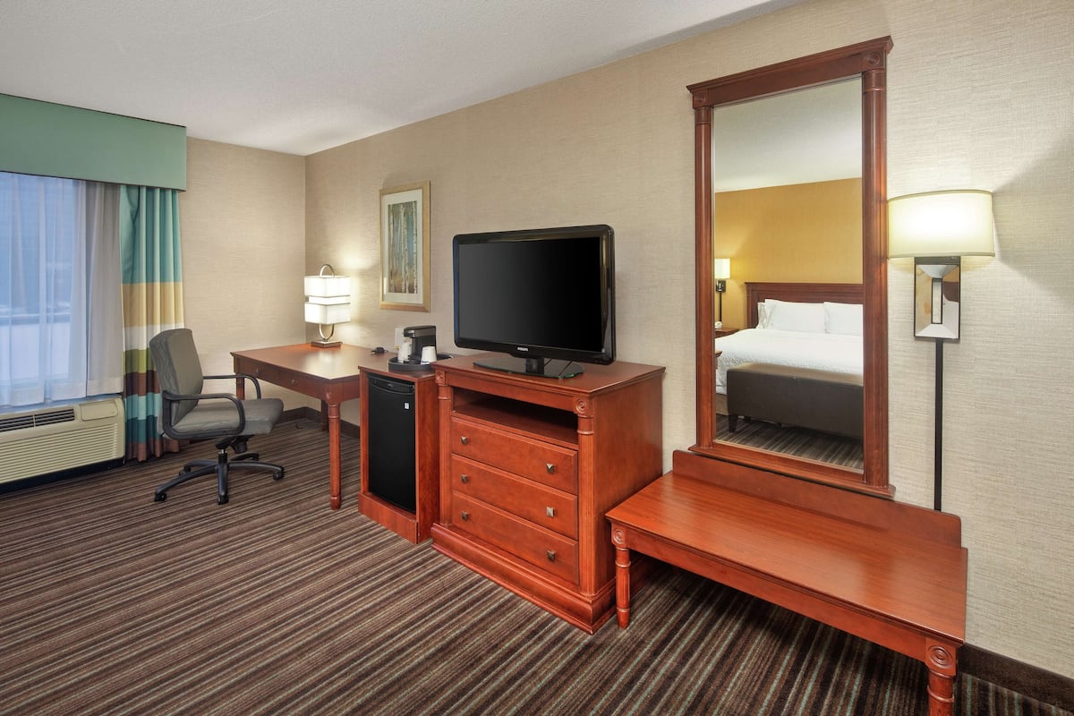 A hotel room with a bed, a desk with a television, a dresser, a chair, and a lamp.