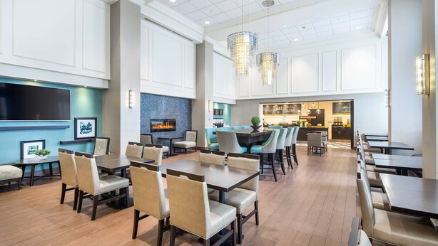 A spacious dining area with multiple tables and chairs, a kitchen in the background, and a flat-screen TV mounted on the wall. - Hampton Inn & Suites by Hilton Toronto Airport (Mississauga)