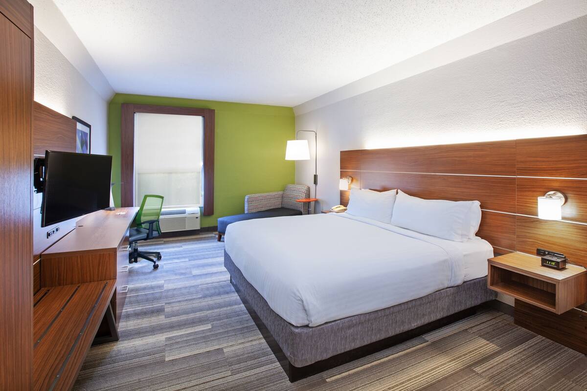 Ein Hotelzimmer mit einem großen Bett, einem Schreibtisch mit Computer, einem Stuhl und einem Fenster mit Jalousien.