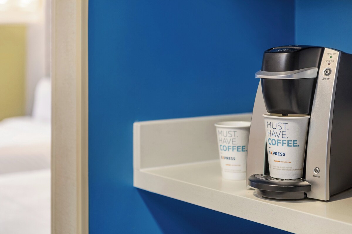 Een koffieapparaat met een kophouder en een koffiekop met de tekst 'MUST. HAVE. COFFEE.' op een witte plank tegen een blauwe muur.