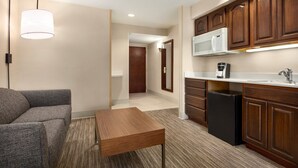 A modern kitchen with wooden cabinets, a microwave, and a refrigerator. There is a gray sofa and a wooden table in the living area. - Holiday Inn Express & Suites Philadelphia - Mt. Laurel by IHG (Mount Laurel)