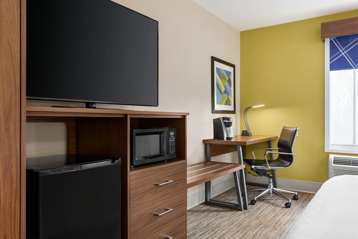 A hotel room with a flat-screen TV, a microwave, a desk with a chair, a framed picture on the wall, and a window with blinds.