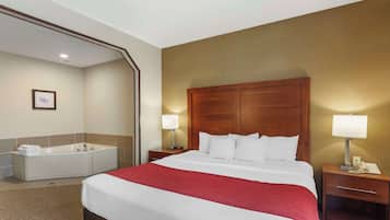 A hotel room with a large bed, a wooden headboard, two bedside tables with lamps, a bathroom with a bathtub, and a framed picture on the wall.