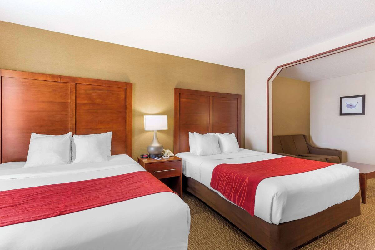 A hotel room with two beds, wooden headboards, white bedding, a red throw blanket, a nightstand with a lamp, a chair, and a framed picture on the wall.