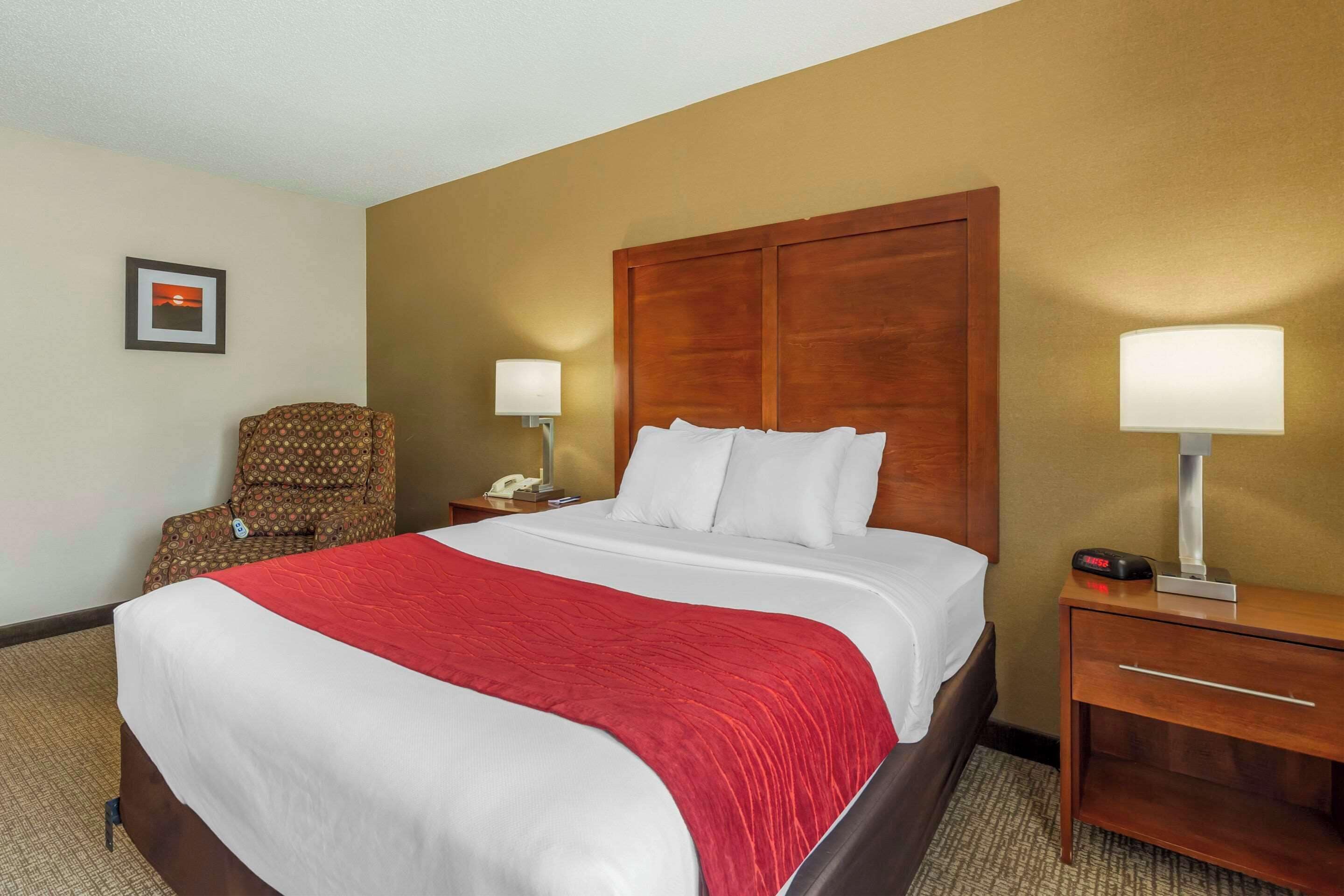 A hotel room with a large bed, a wooden headboard, a red blanket, a chair, a nightstand with a lamp, and a framed picture on the wall.