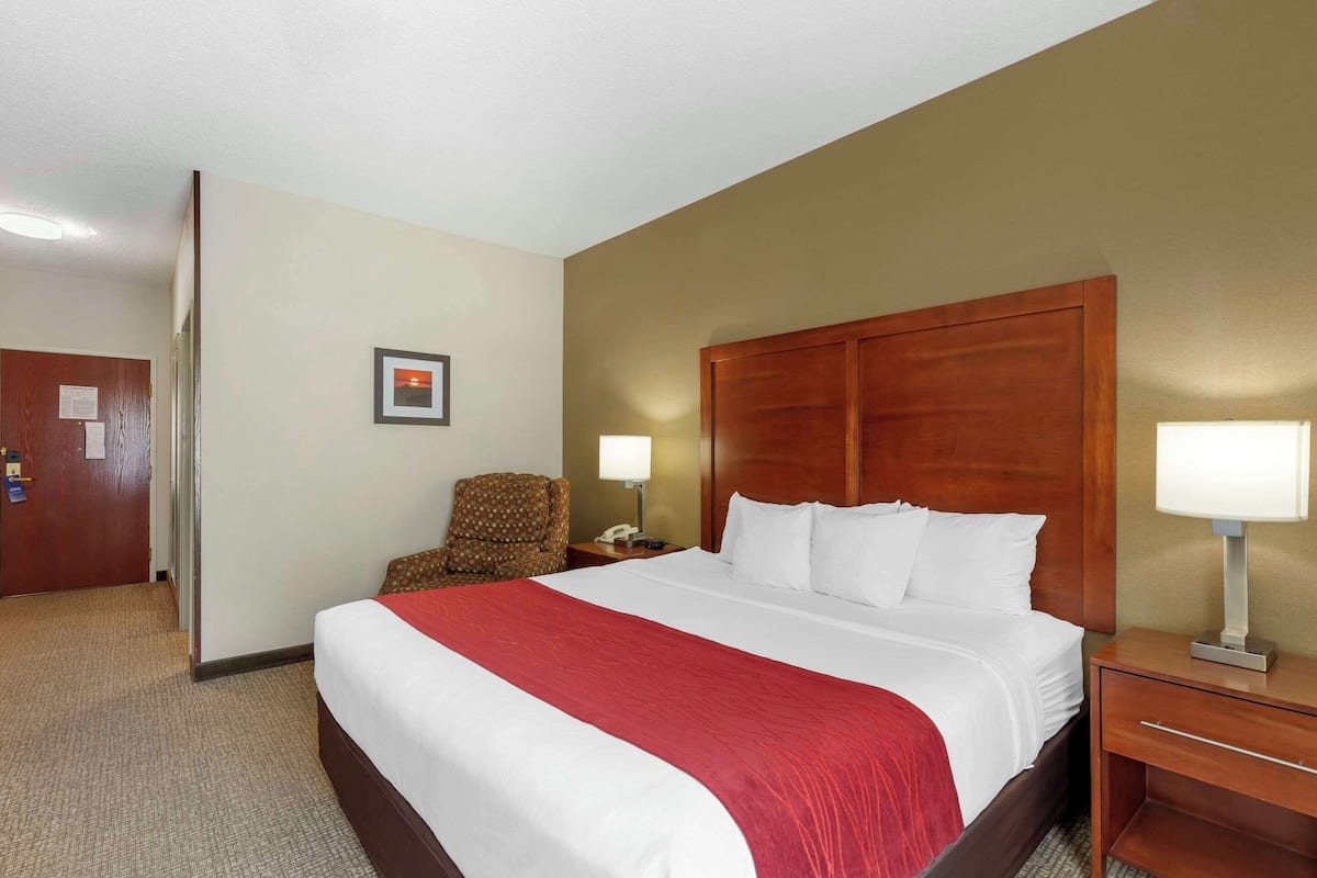 A hotel room with a large bed, a wooden headboard, a nightstand with a lamp, a chair, and a door with a sign.