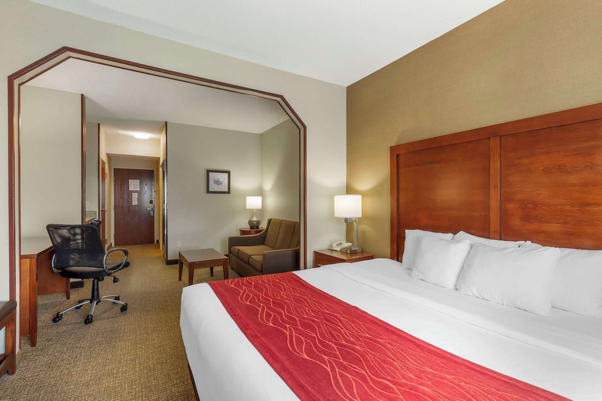 A hotel room with a large bed, a desk with a chair, a small table with a lamp, and a mirror.