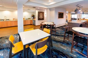 Hall d'hotel con zona pranzo centrale, caratterizzata da tavoli rotondi e sedie gialle, angolo cucina e un bar.