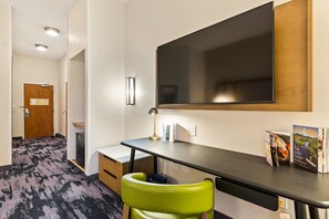 A hotel room with a desk, a green chair, a flat-screen TV, a lamp, and a magazine on the desk. - Fairfield Inn & Suites by Marriott Bend Downtown (Bend)