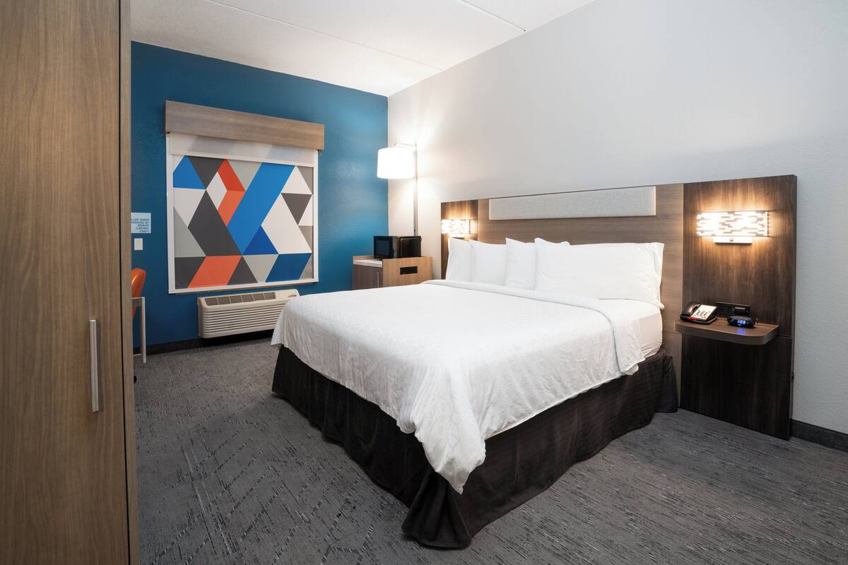 A hotel room with a large bed, a bedside table with a lamp, a small desk, and a wall-mounted TV.