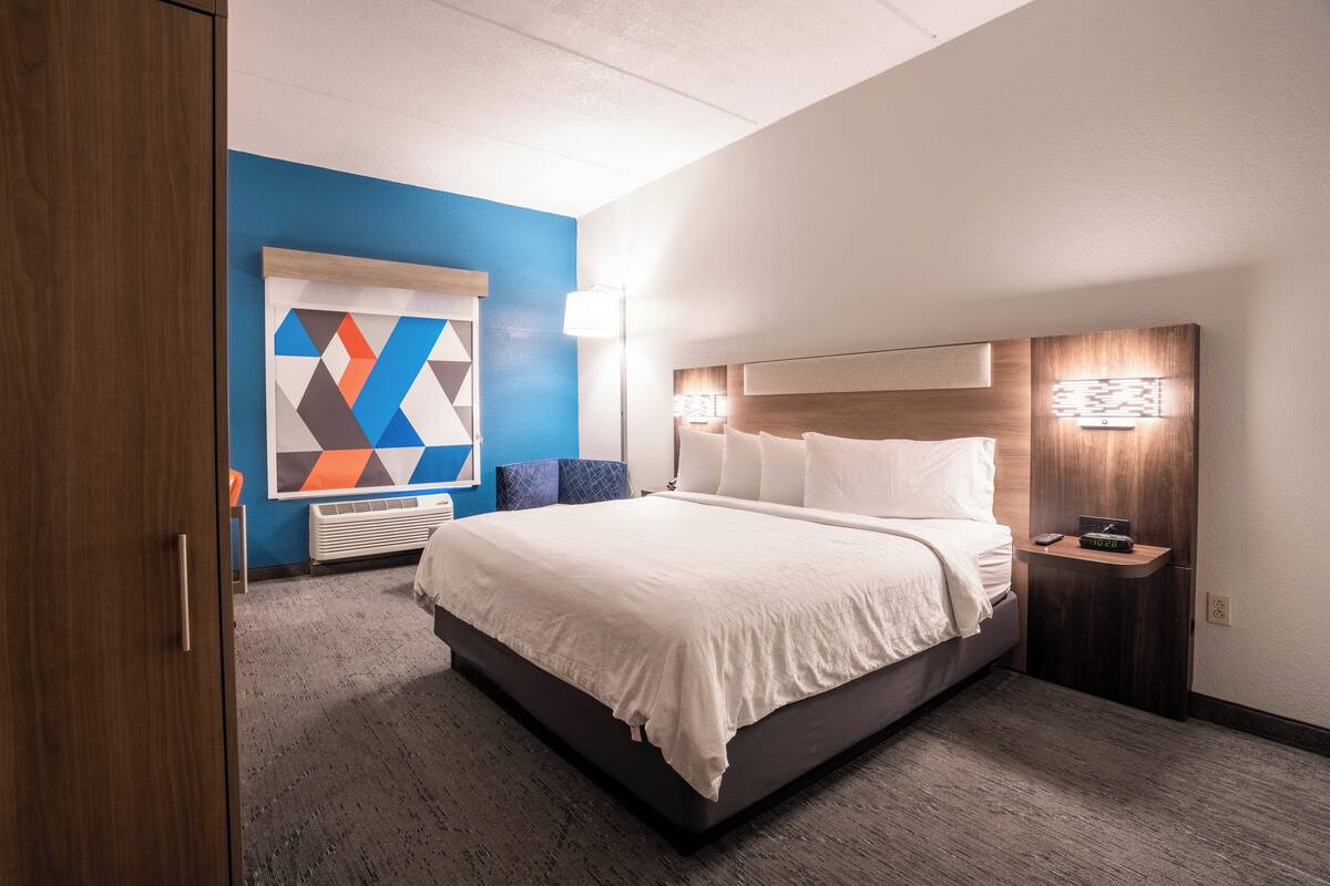 A hotel room with a large bed, a nightstand with a lamp, a wall-mounted TV, and a colorful abstract painting on the wall.