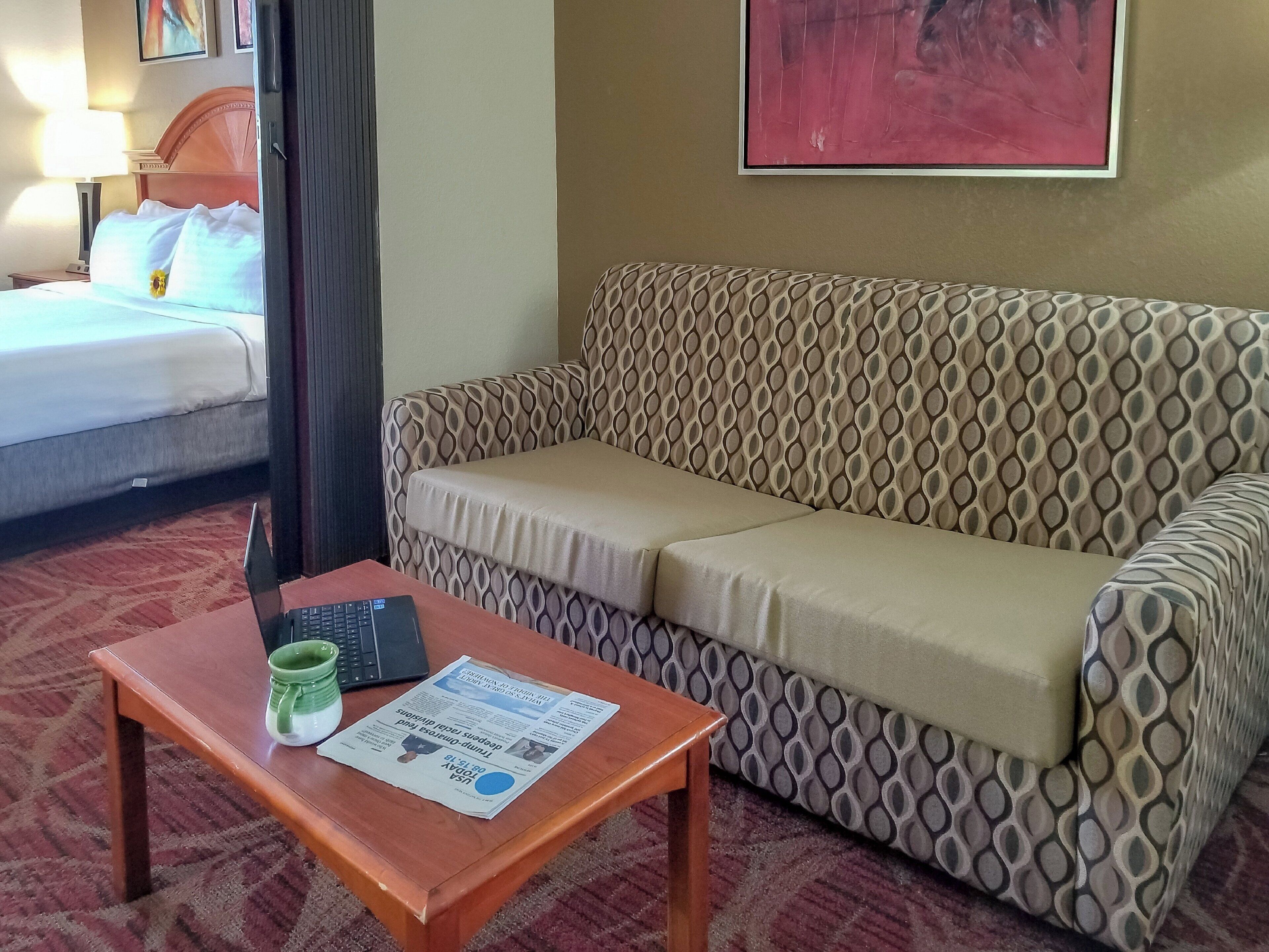 A hotel room with a bed, a patterned sofa, a small wooden table with a laptop and mug, and a framed picture on the wall.