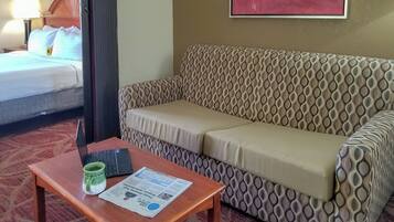 A hotel room with a bed, a patterned sofa, a small wooden table with a laptop and mug, and a framed picture on the wall.