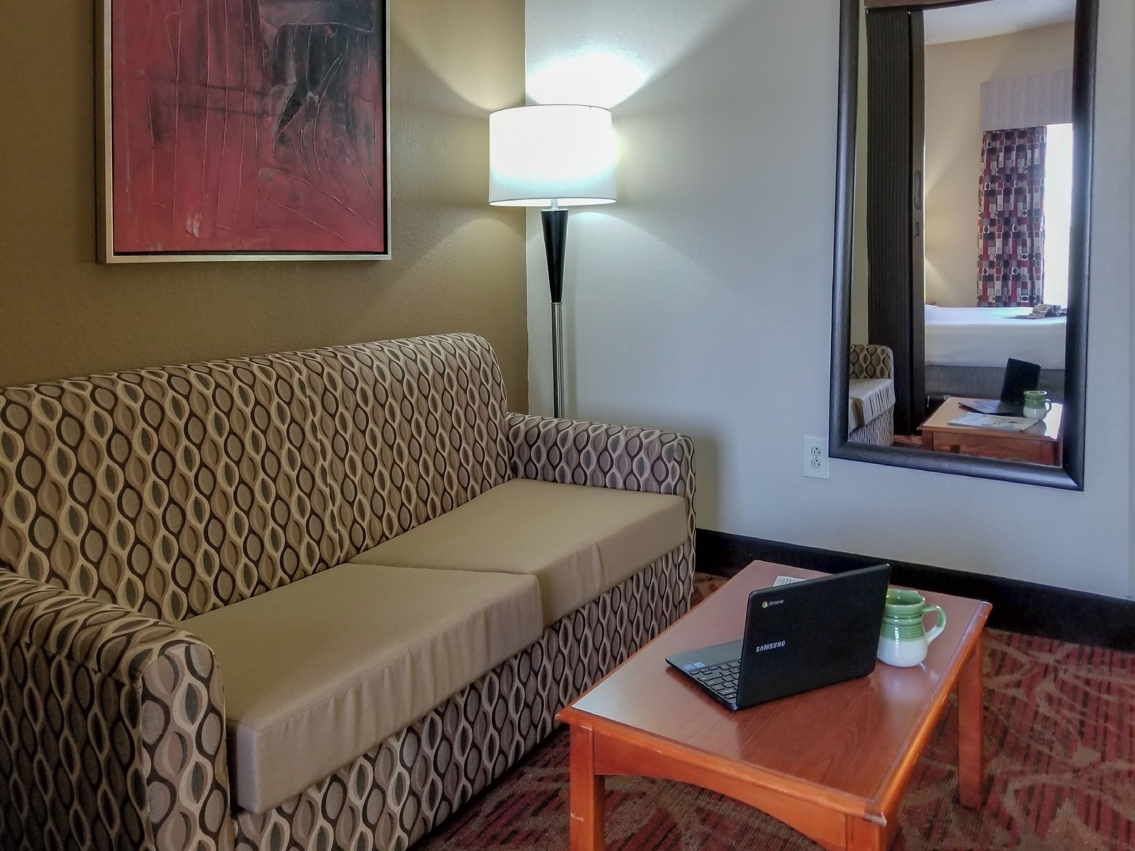 Ein Hotelzimmer mit einer gemusterten Couch, einer Lampe, einem Laptop auf einem kleinen Tisch und einem Spiegel, der den Raum reflektiert.