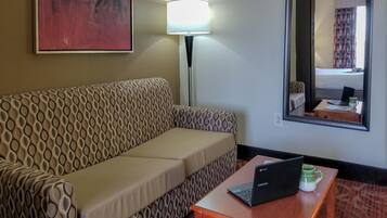 Ein Hotelzimmer mit einer gemusterten Couch, einer Lampe, einem Laptop auf einem kleinen Tisch und einem Spiegel, der den Raum reflektiert.