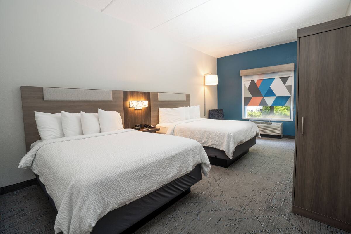 A hotel room with two beds, a nightstand, a wall-mounted lamp, and a window with a geometric view.