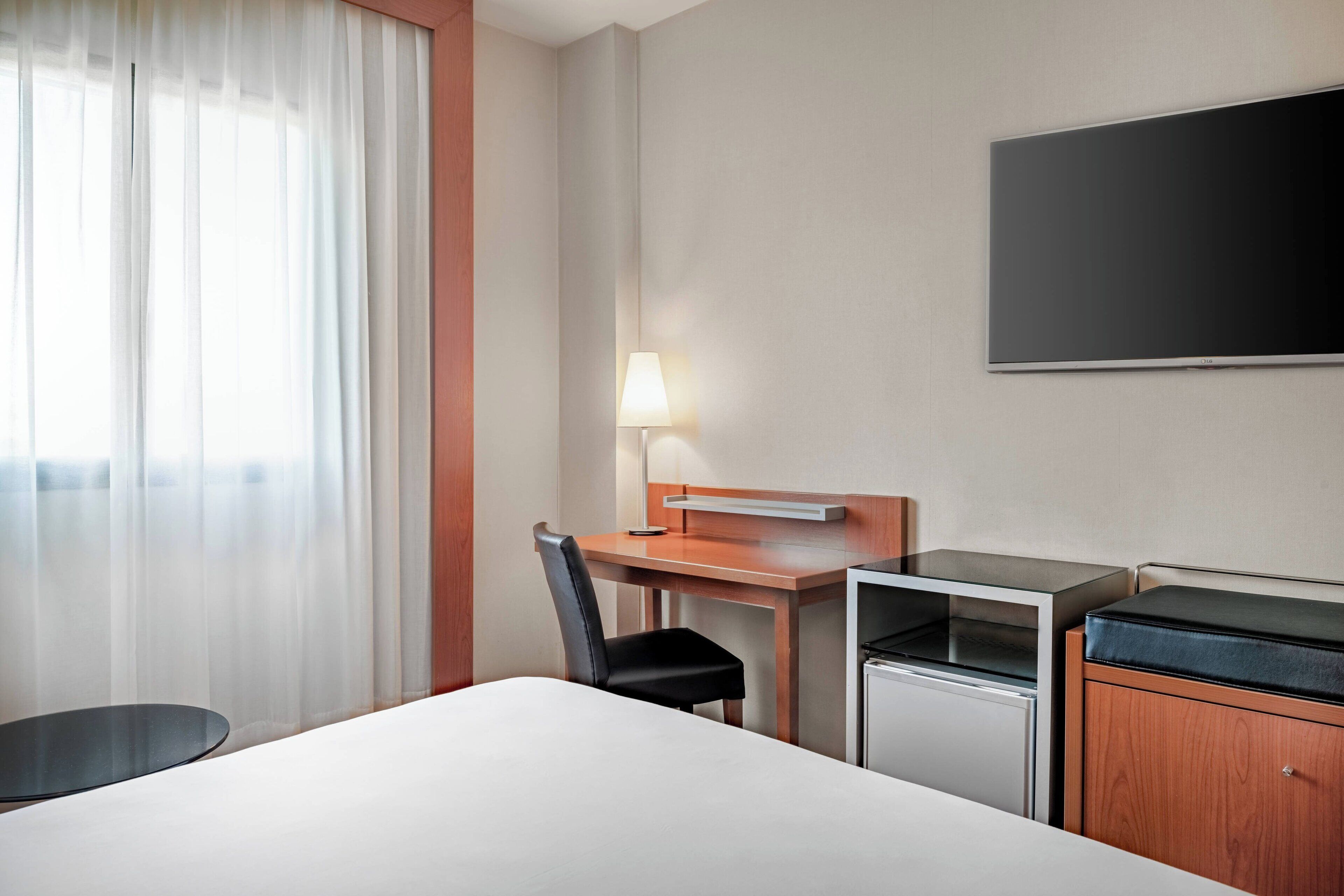 Una habitación de hotel con cama, escritorio y silla, lámpara, televisor y minibar.