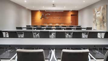 A conference room with a long table and chairs, a wooden wall with "AC HOTELS" signage, and a projector on the ceiling.